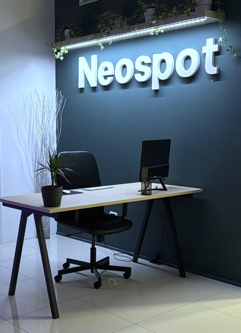 Una postazione di lavoro nell’ufficio Neospot, con l’insegna sulla parete e le piante accanto alla scrivania